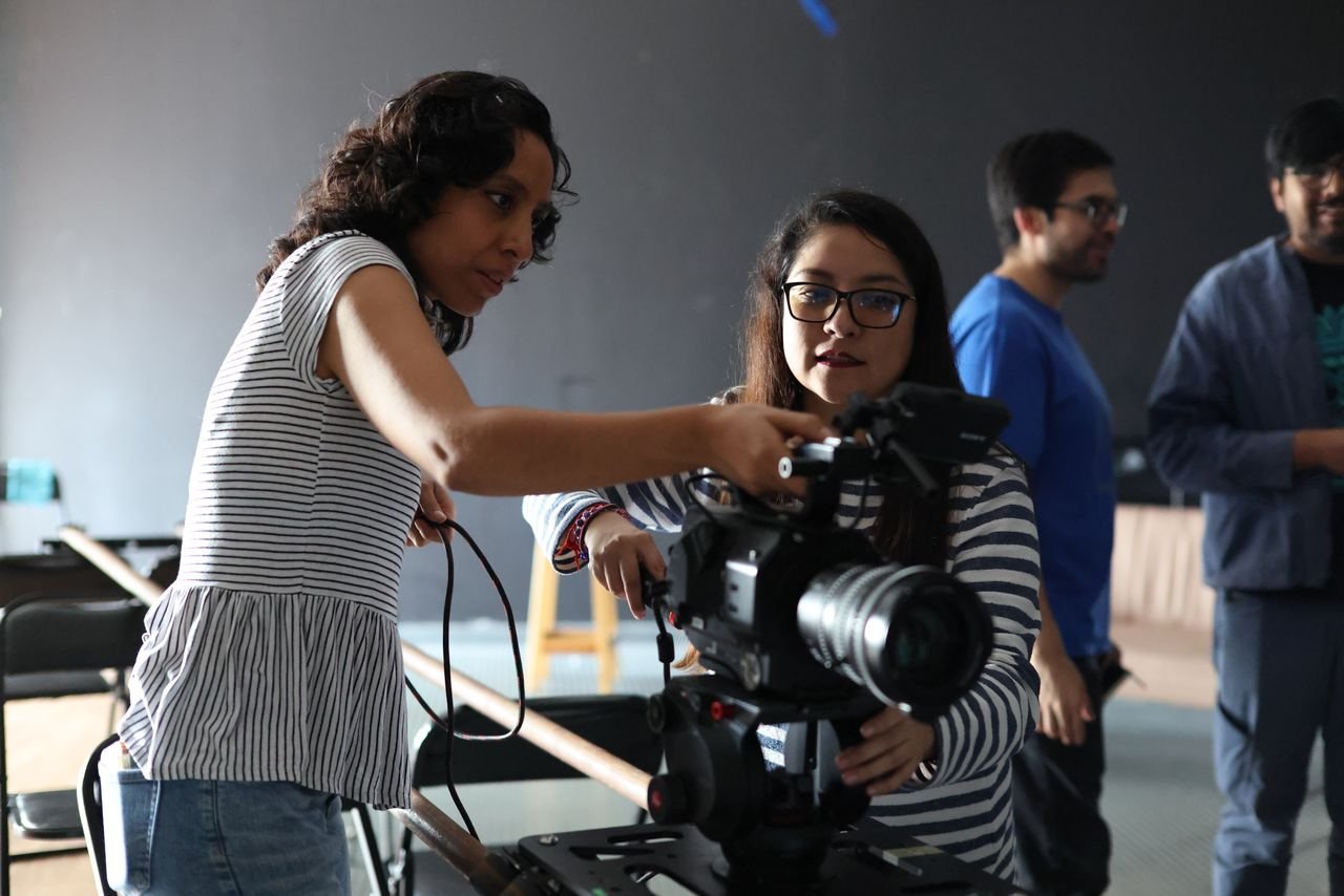 Two women work together on a film set, adjusting camera equipment indoors.